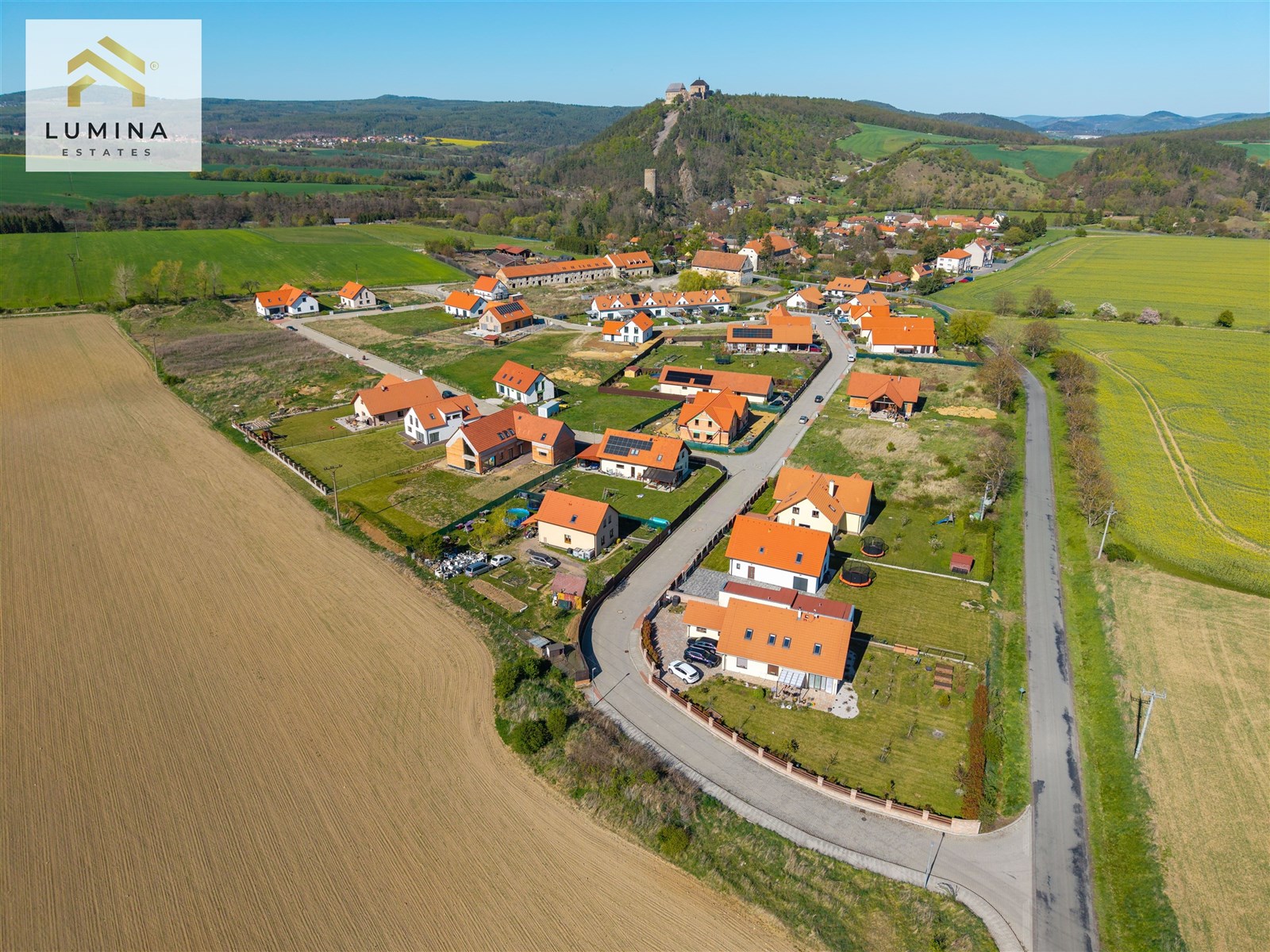 Letecký pohled na novou rezidenční čtvrť s moderními domy s červenými střechami, obklopenou zelenými poli a malebnou krajinou.
