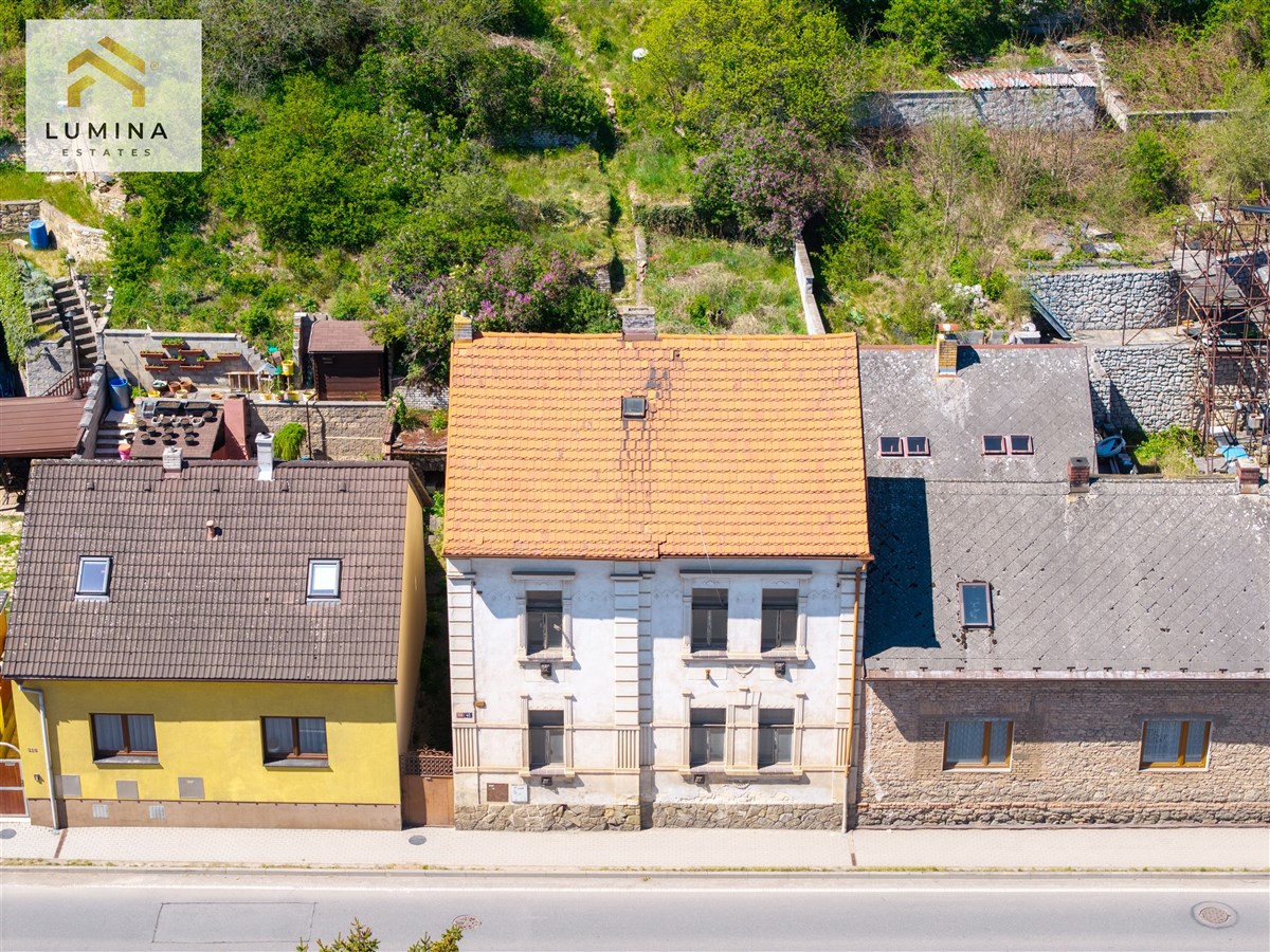 Starší rodinný dům s červenou střechou, vedle dalších dvou domů, zasazený v zelené zahradě. Potenciál pro rekonstrukci a renovaci.