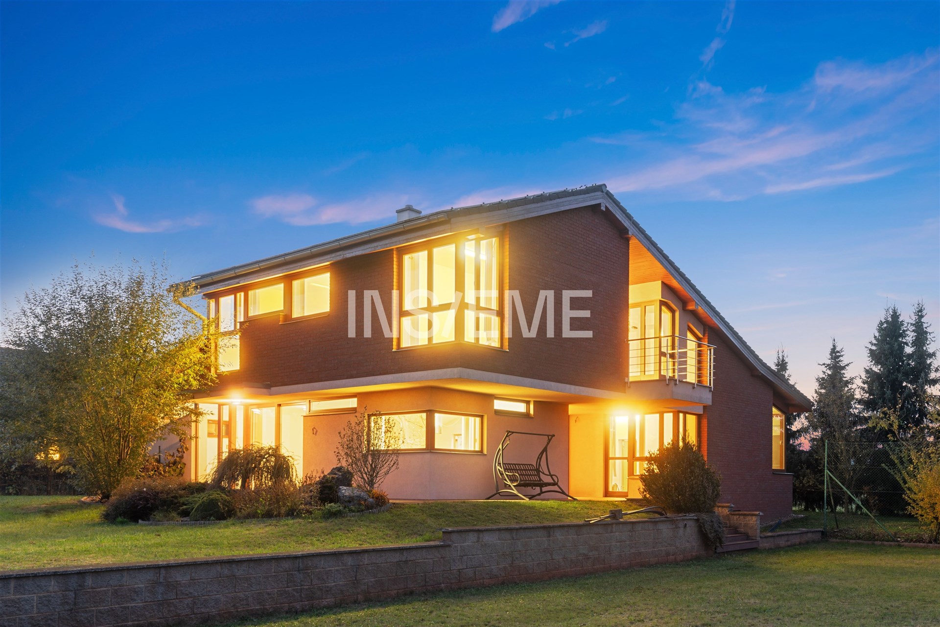 Moderní rodinný dům s cihlovou fasádou a velkými okny, prostorná zahrada a osvětlené interiéry vytvářejí elegantní a útulný vzhled