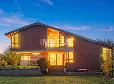 Dvoupodlažní rodinný dům s cihlovou fasádou a velkými okny, moderní design, upravená zahrada, večerní osvětlení vytváří útulnou atmosféru. | Prodej - dům/vila, 329 m²