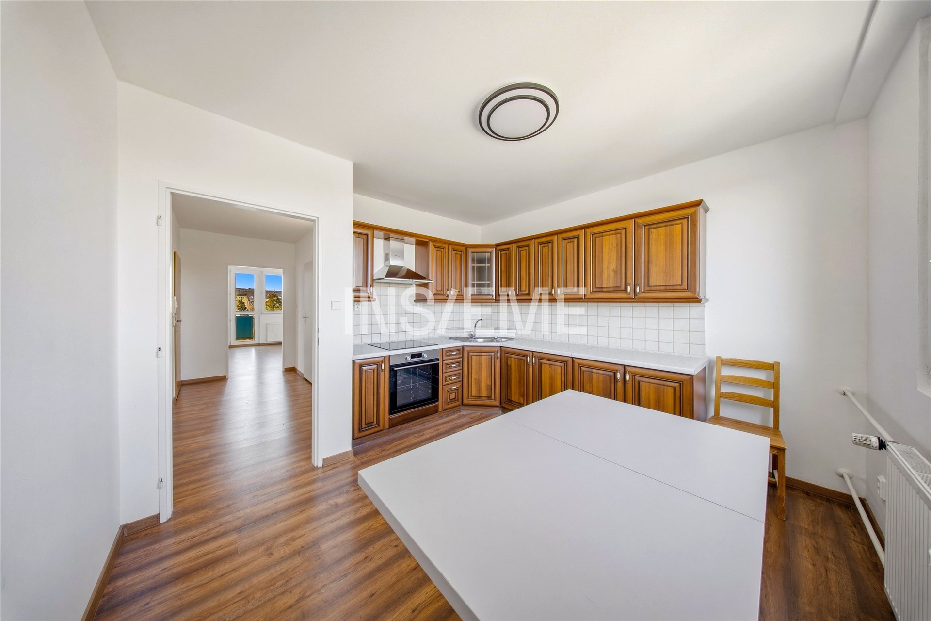 Prostorná kuchyň s dřevěnými skříňkami, moderními spotřebiči a parketovou podlahou, vedlejší světlý obývací pokoj s velkým oknem.