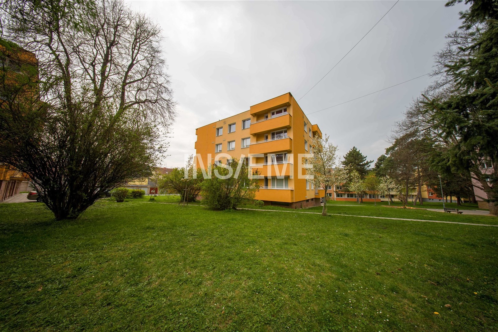 Čtyřpodlažní bytový dům s barevnou fasádou obklopený zelení, v tiché a klidné lokalitě, s udržovaným trávníkem a blízkostí stromů.