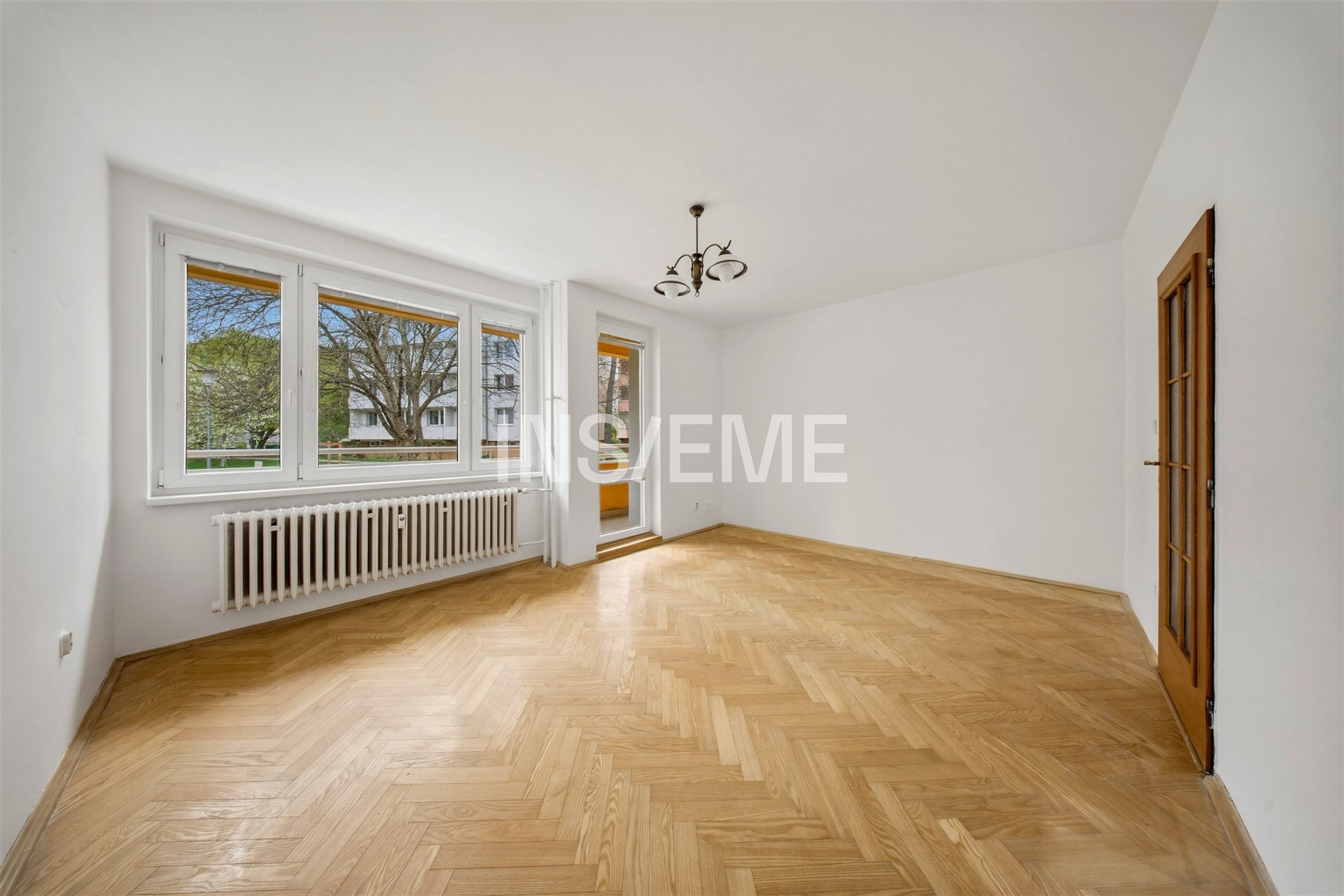 Světlý obývací pokoj s velkými okny, parketovou podlahou a moderním osvětlením, nabízí výhled do zahrady a vstup na balkon.