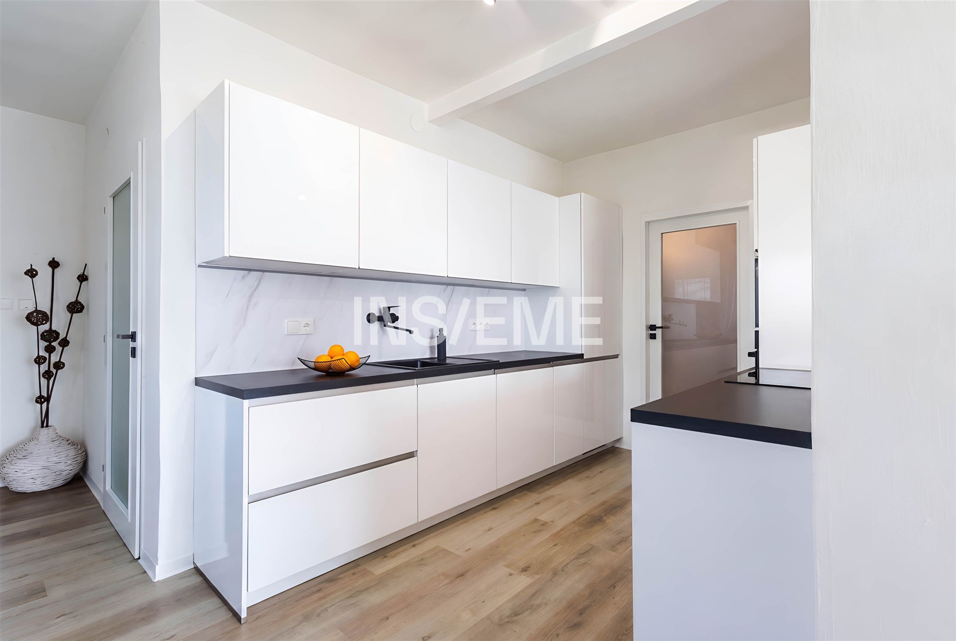 Moderní kuchyň s bílými skříňkami, černou pracovní deskou a parketovou podlahou, prostorná a světlá s minimalistickým designem.