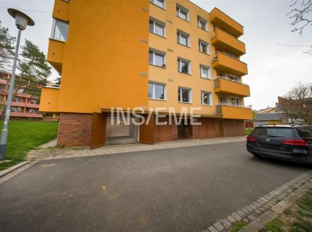 Vchod do bytového domu s moderní fasádou a nezastřešeným parkováním, obklopený zelení a přilehlou asfaltovou cestou pro parkování. | Prodej - malý objekt/garáž