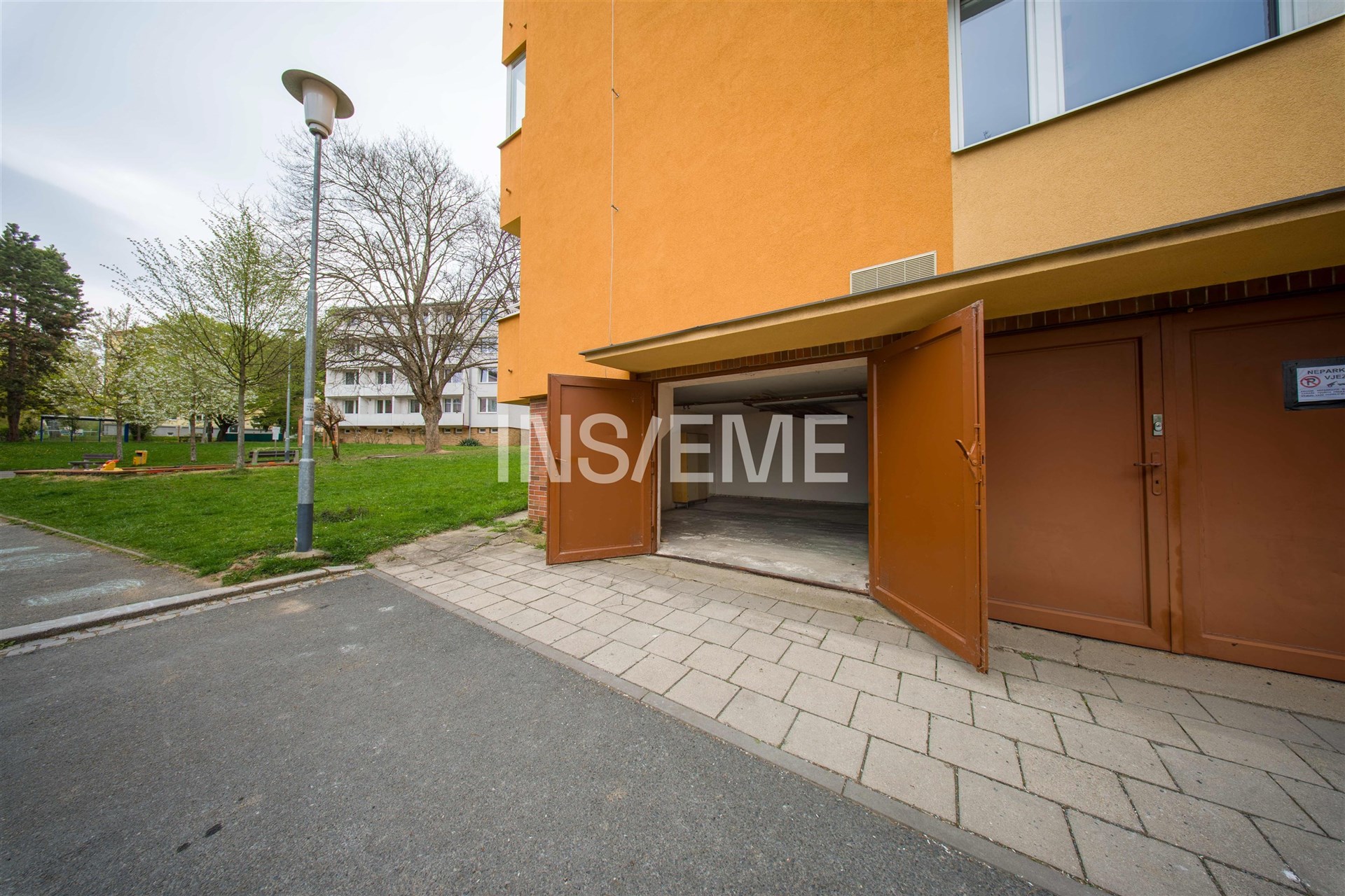 Otevřená garáž s betonovou podlahou a oranžovou fasádou, vedle zeleného parku, nabízí praktický úložný prostor v klidné lokalitě.