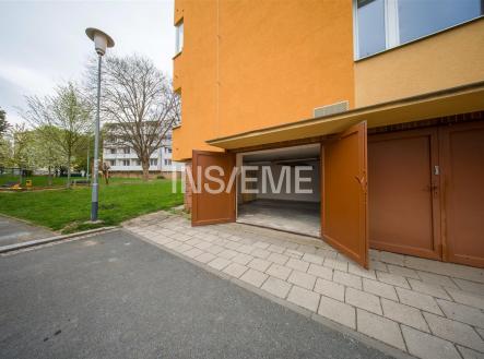 Otevřená garáž s betonovou podlahou a oranžovou fasádou, vedle zeleného parku, nabízí praktický úložný prostor v klidné lokalitě. | Prodej - malý objekt/garáž