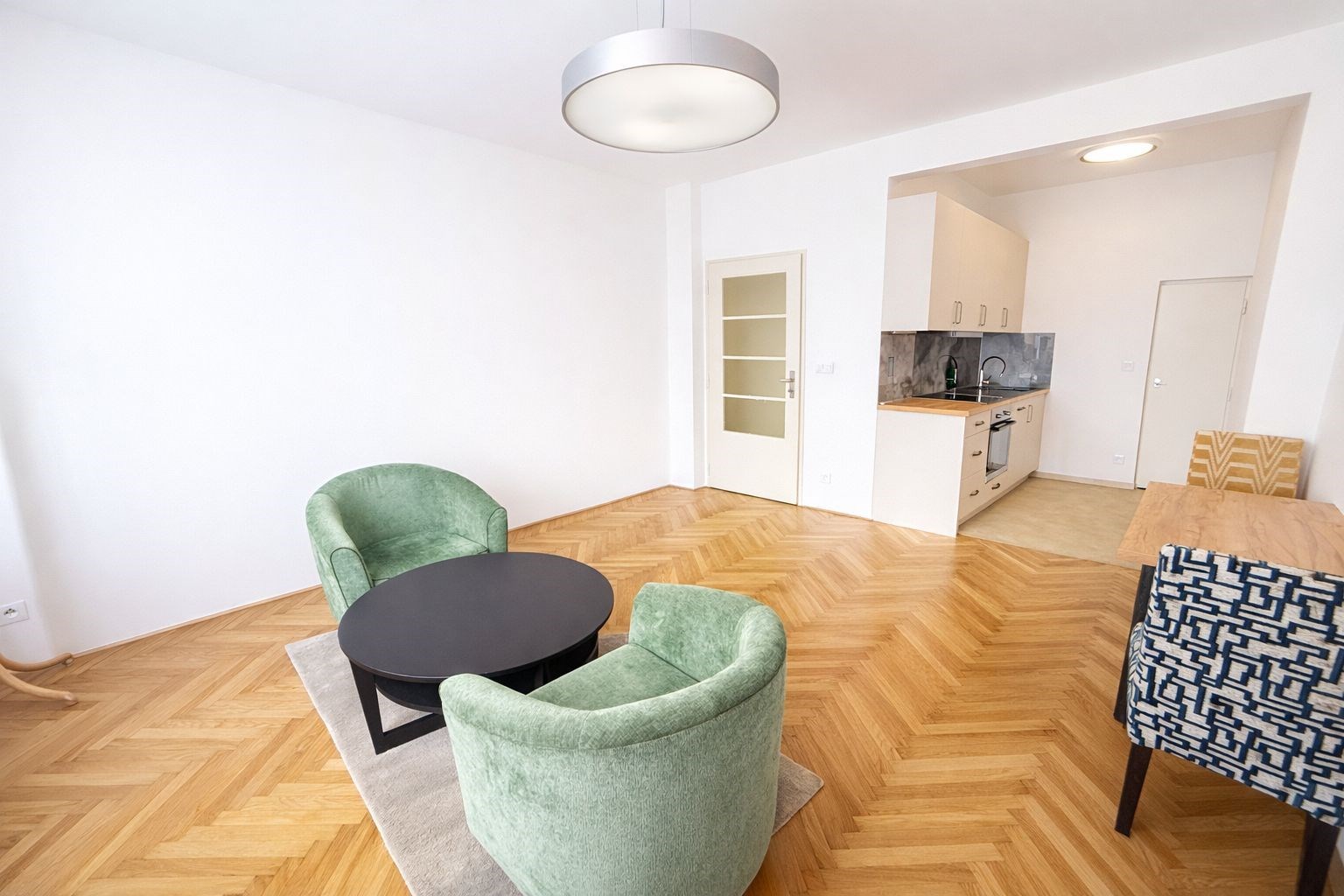 Světlý obývací pokoj s parketovou podlahou, moderní kuchyňskou linkou a velkými okny. Prostor je světlý a přehledný.