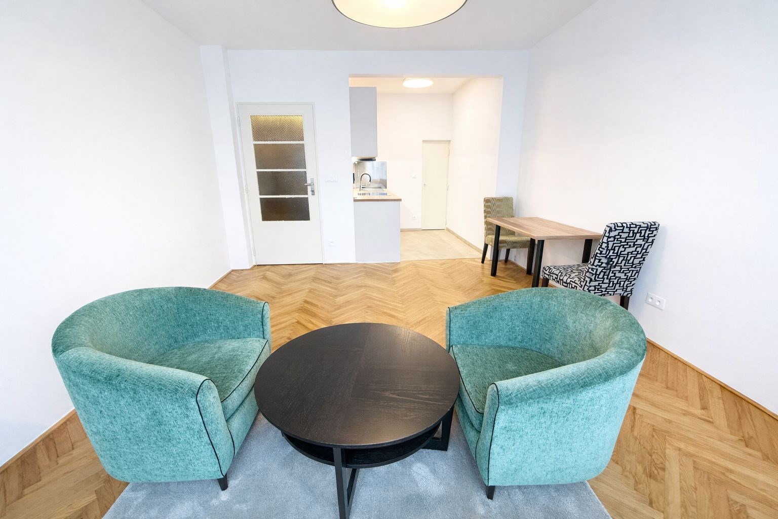 Světlý obývací pokoj s parketovou podlahou, moderním nábytkem a přístupem do otevřené kuchyně. Minimalistický design a velká okna