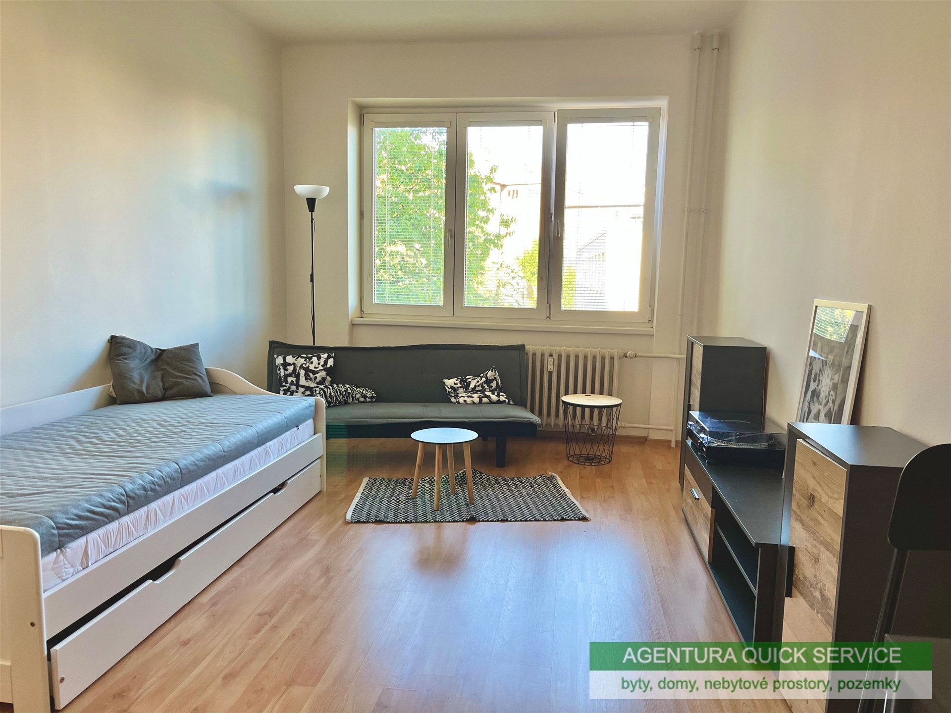 Světlý obývací pokoj s parketovou podlahou, velká okna zajišťují dostatek přirozeného světla, moderní minimalistický nábytek.