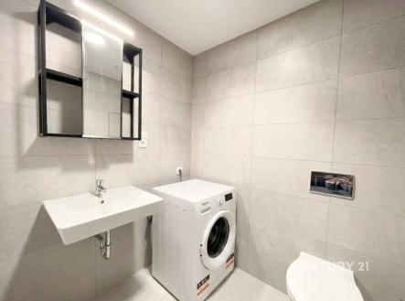 Moderní koupelna s dlažbou, která zahrnuje zrcadlovou skříňku a pračku, nabízí praktické a čisté řešení s moderním designem. | Pronájem bytu, garsoniéra, 36 m²