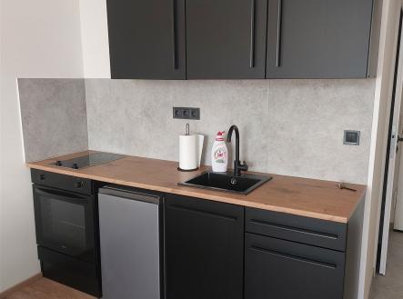 Moderní kuchyň s dřevěnou pracovní deskou, černými skříňkami a parketovou podlahou vytváří elegantní a funkční prostor. | Pronájem bytu, 1+kk, 30 m²