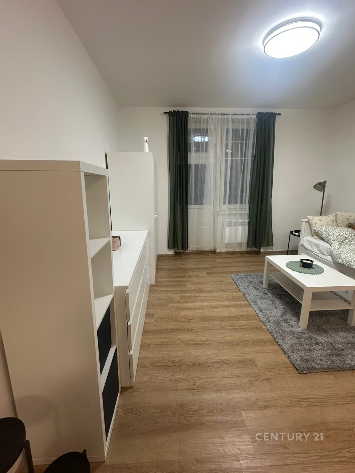 Světlý obývací pokoj s parketovou podlahou, moderním osvětlením a velkými okny, jednoduchý a útulný styl interiéru.