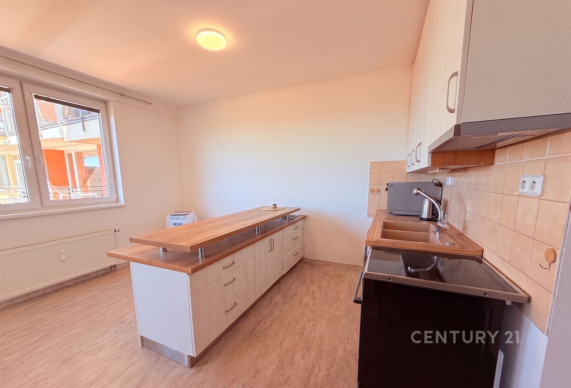 Kuchyňský kout s moderními skříňkami, světlou dřevěnou podlahou a velkými okny, které poskytují dostatek přirozeného světla