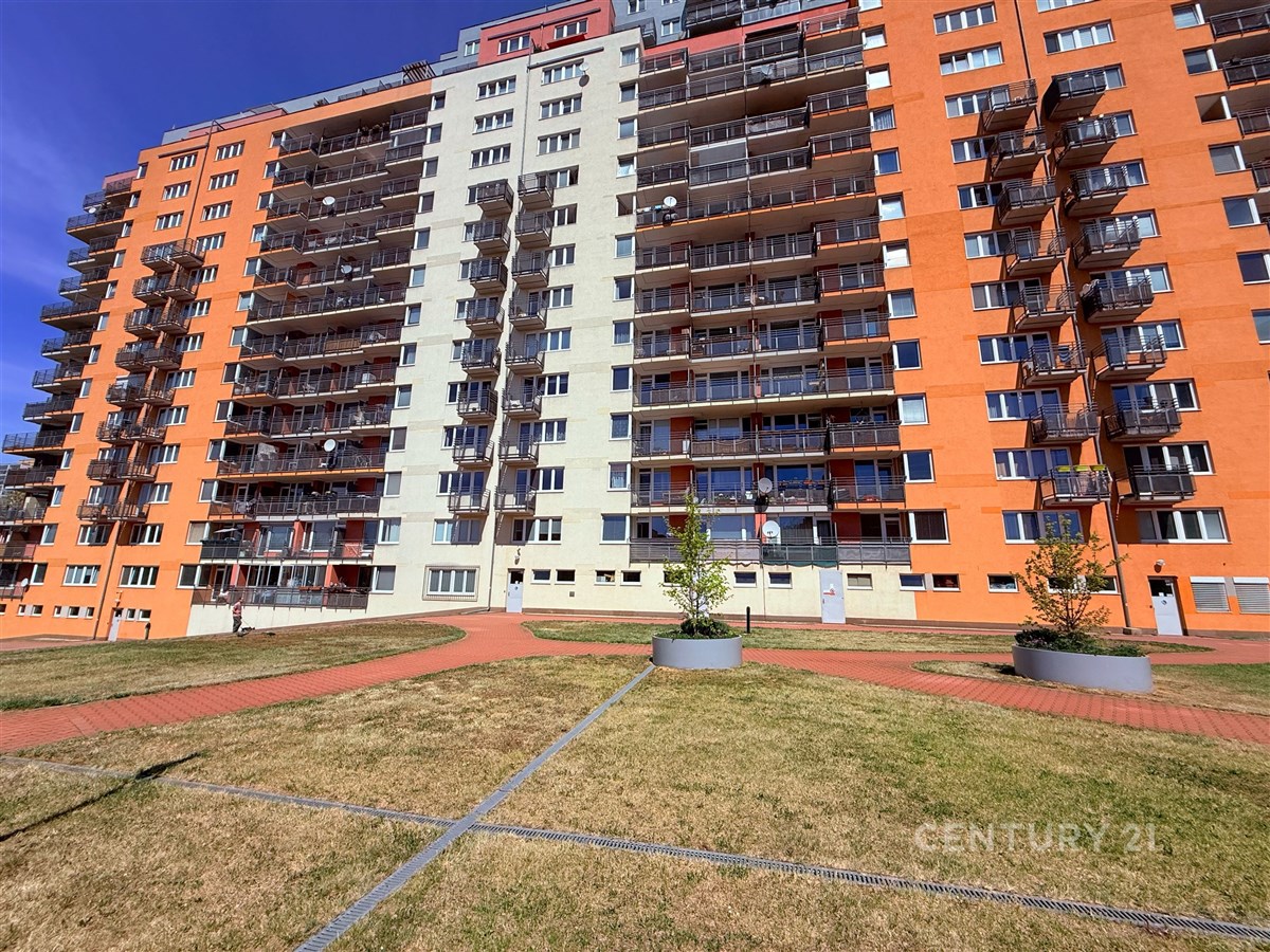 Výšková bytová budova s balkony, moderní fasáda v oranžové a světle šedé barvě, udržovaný trávník a dlážděné chodníky v okolí