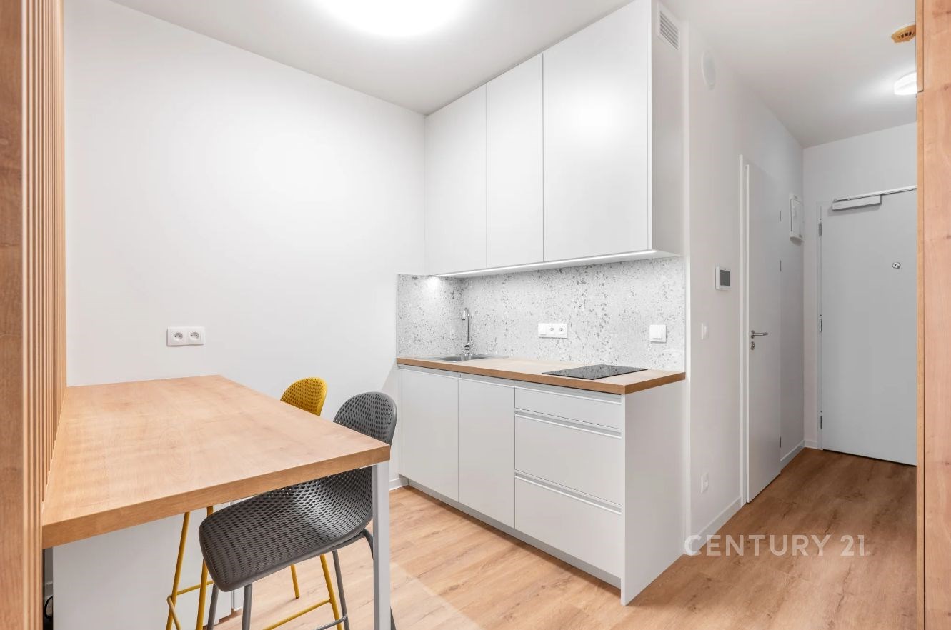 Kompaktní kuchyň s moderním nábytkem, bílými skříňkami a dřevěnou podlahou, doplněná světlým obkladem a jídelním koutem pro dva.