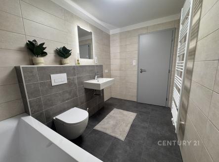 Moderní koupelna s dlažbou, světlými obklady a závěsnou toaletou, vybavená umyvadlem a velkým zrcadlem, nabízí elegantní design. | Pronájem bytu, 1+kk, 39 m²