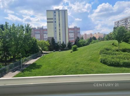 Výhled z okna na upravený zelený park v sídlišti s panelovými domy, modrá obloha, klidná lokalita, blízkost přírody a městské infrastruktury | Pronájem bytu, 2+kk, 60 m²