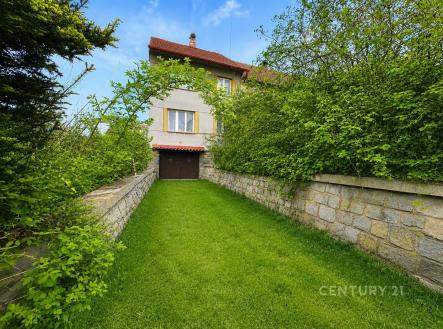 Zahrada s upraveným trávníkem a kamennou zdí u rodinného domu. Dům má moderní fasádu, červenou střechu a je obklopen zelení. | Prodej - dům/vila, 500 m²