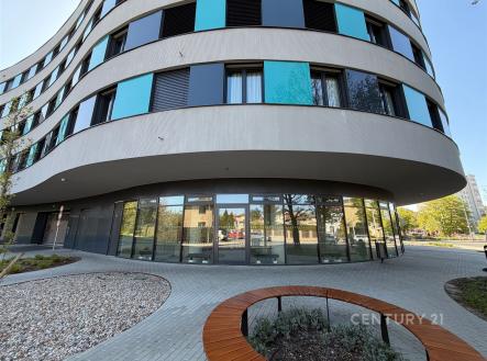 Moderní bytový dům s velkými okny a elegantní fasádou, obklopený dlažbou a upravenými venkovními prostory v klidné části města. | Pronájem - obchodní prostor, 84 m²